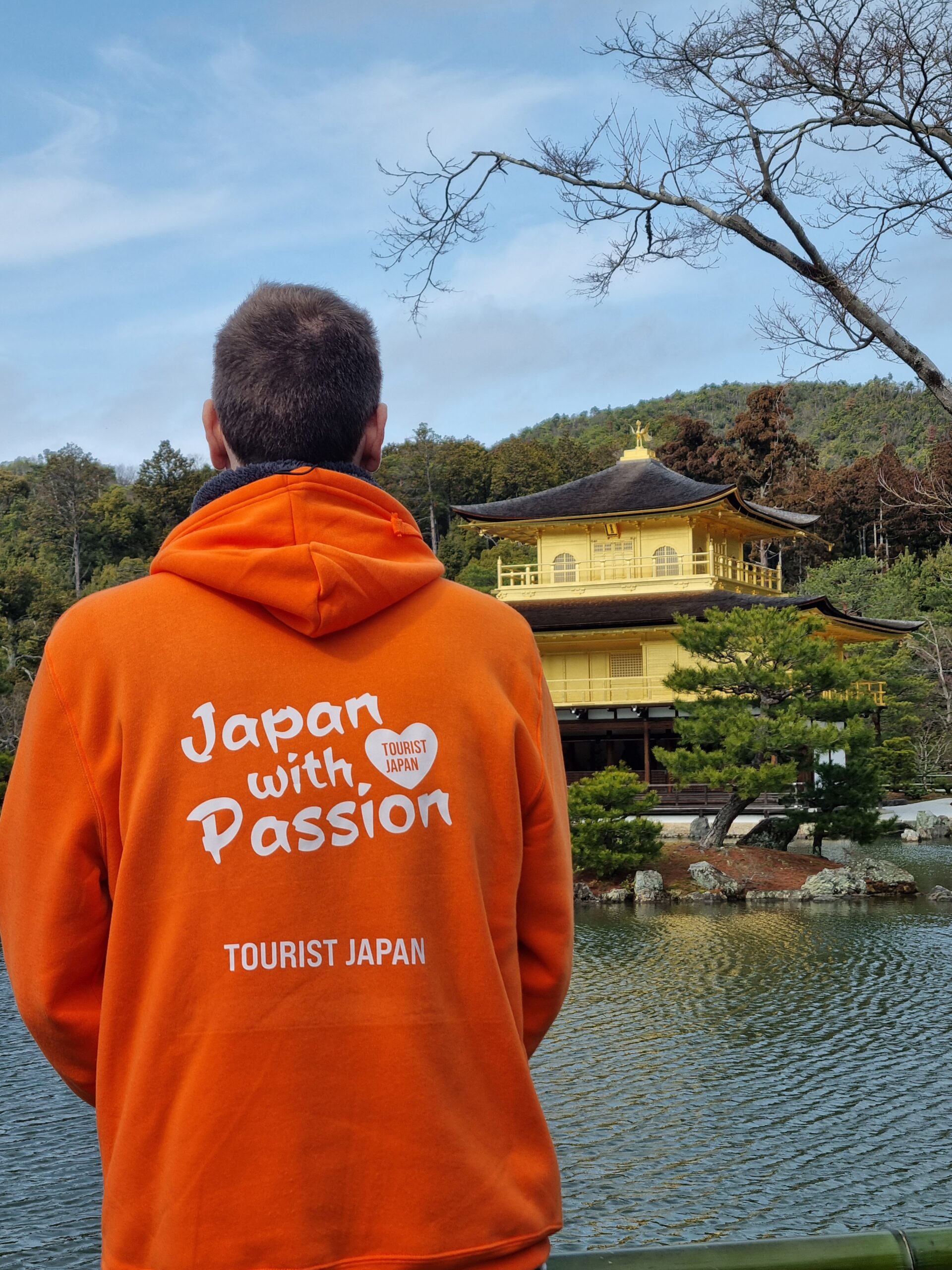 Young Man In An Orange Hoodie With Tourist Japan Branding And The Tagline Japan With Passion, Looking Out At The Golden Pavillion In Kyoto